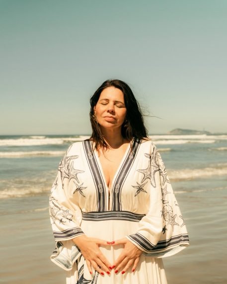 Cami Loureiro em um momento de presença, com as mãos sobre o ventre, à beira-mar